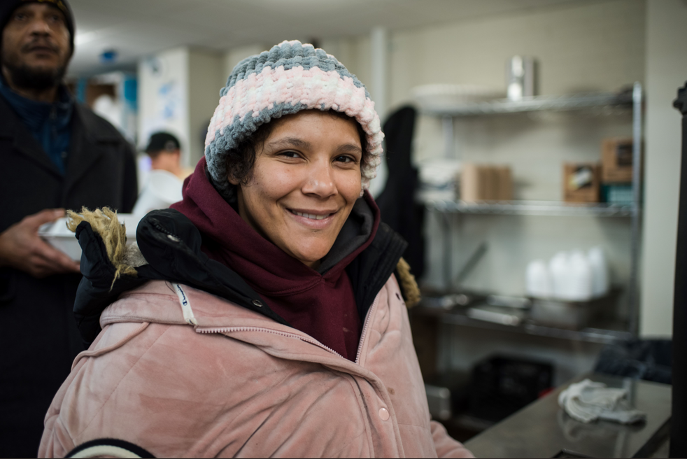A St. Francis House guest wearing a knit beanie and pink coat smiles at the camera, standing in the dining area.